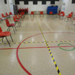 A large main hall / basketball court, with social distancing markings laid out on the floor. Stackable chairs are visible around the perimeter as are some tables and crash-mats.